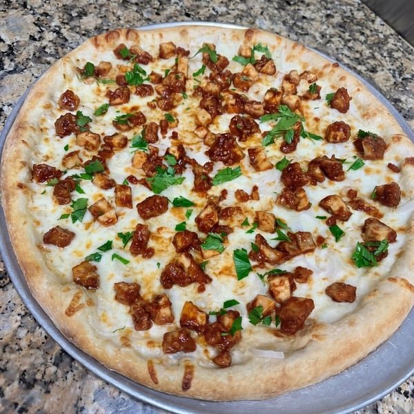 A whole BBQ chicken pizza with white sauce, mozzarella, chicken pieces, and fresh parsley on a metal pan.