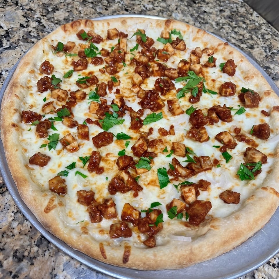 A whole BBQ chicken pizza with white sauce, mozzarella, chicken pieces, and fresh parsley on a metal pan.