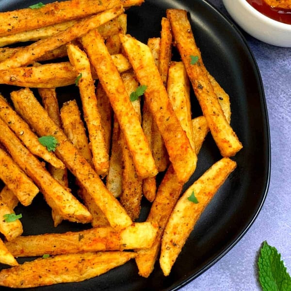 Golden-brown seasoned fries with herbs on a black plate, with a small bowl of sauce in the background.