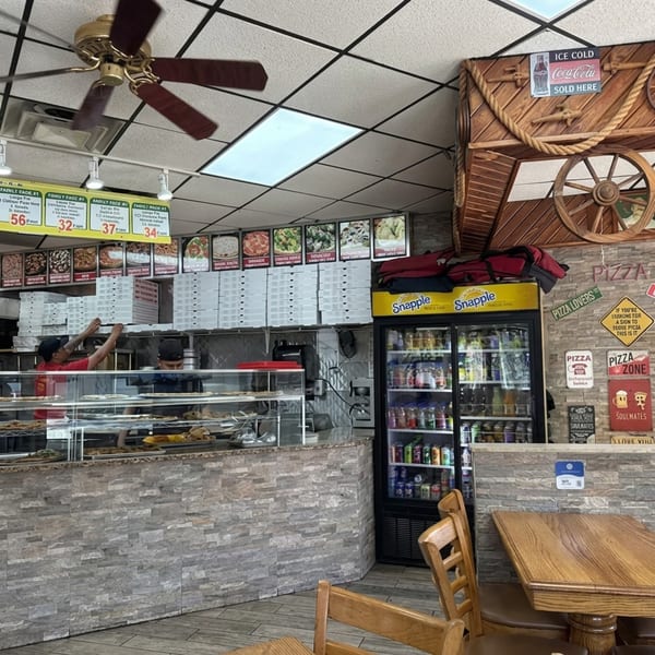 Two employees prepare food behind a counter with pizza boxes, a drink fridge, and wooden tables.